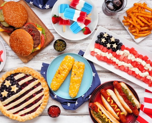 Fourth of July, American food. Above view table scene on white wood. Fourth of July picnic, hamburgers, hotdogs, blueberry pie, red, white, blue popsicles, American flag made out of fruit