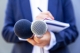 man in blue suit holding pen, notebook and 2 microphones; rebrand press release