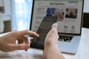Man Reading News Online On A Smartphone And Laptop. Reads Articles, Newspaper, Information Concept hands holding smartphone in front of open laptop; current events September 2025; Iran conflict surging oil prices