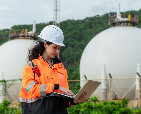 Focused female engineer working on a laptop, conducting a systems check at a natural gas facility. Woman technician analyzing data on a computer in front of large industrial spherical tanks.