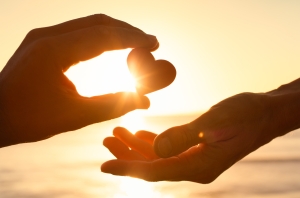 Hand giving heart love help support kindness one hand passing a heart to another hand in front of a setting sun; giving thanks