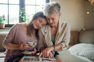 Senior mother with adult daughter indoors at home, looking at family photographs. adult daughter and mother looking at photo album together; Memorial Day honor by remembering