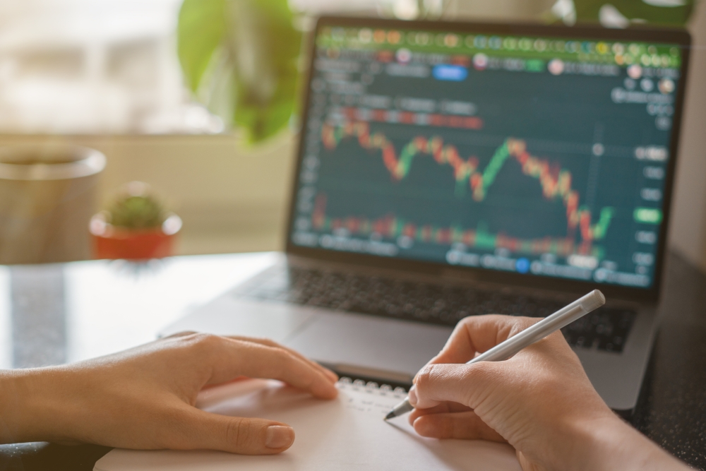 Female hands writing numbers from laptop. Investment analysis concept market graph on screen; hands making notes on pad with a desk