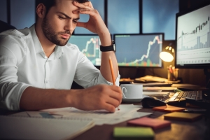 Serious businessman thinking of problem solution working late in office man looking perplexed sitting at desk with monitors with stock info surrounding him